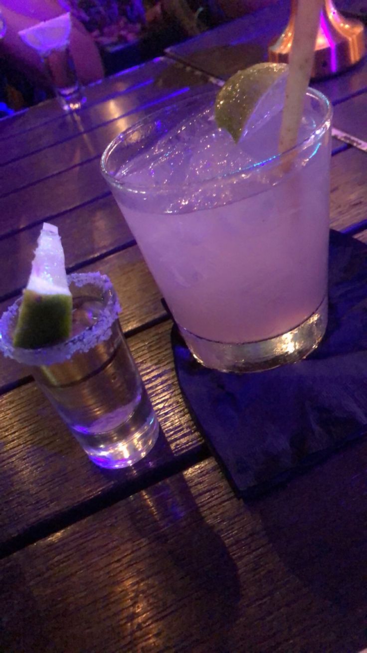 two cocktails are sitting on a table with purple lights in the backround