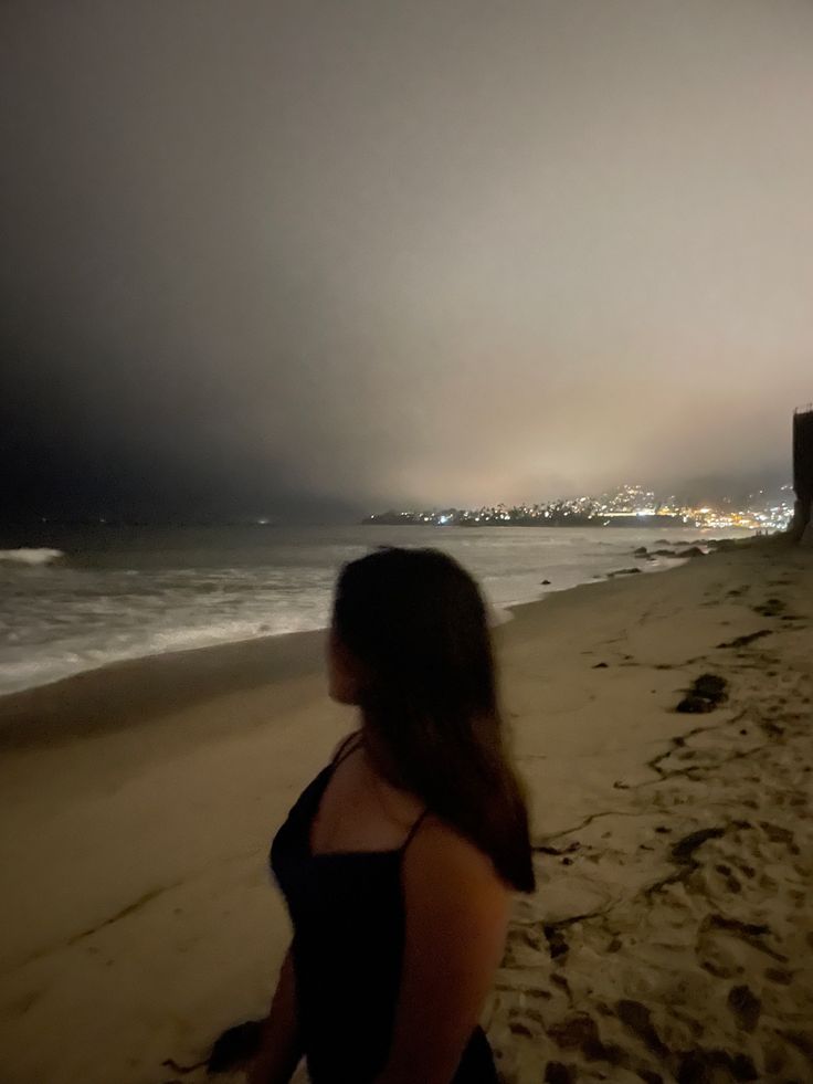 a woman standing on top of a sandy beach next to the ocean under a cloudy sky