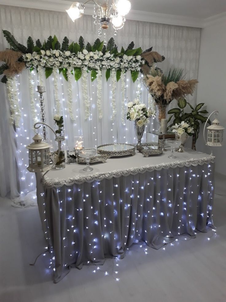 a table covered in white flowers and greenery next to a window with lights on it