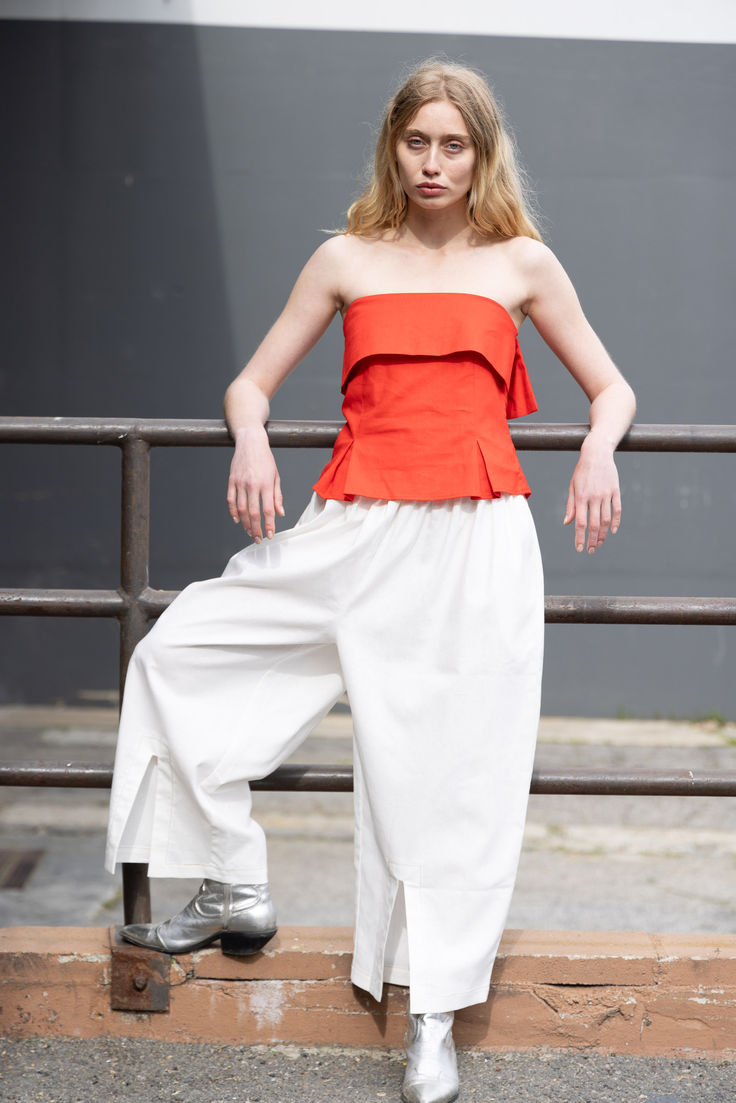 a woman in an orange top and white pants poses for the camera with her hands on her hips