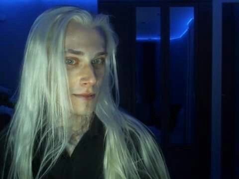 a man with long white hair and blue eyes wearing a black shirt in a dark room