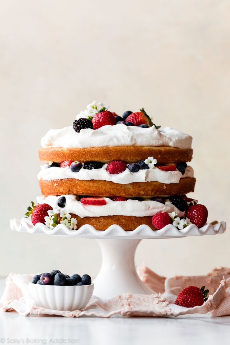 a triple layer cake with berries and whipped cream on top is sitting on a white pedestal