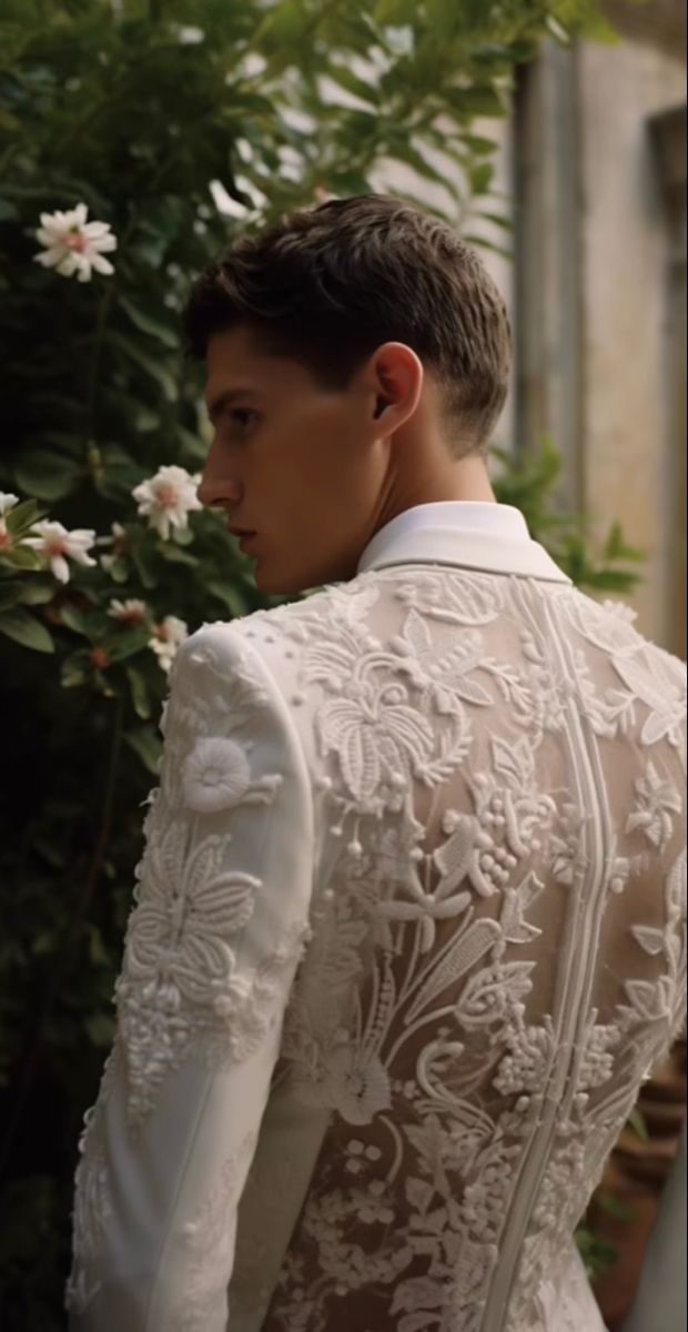 a man in a white suit and tie standing next to a bush with flowers on it