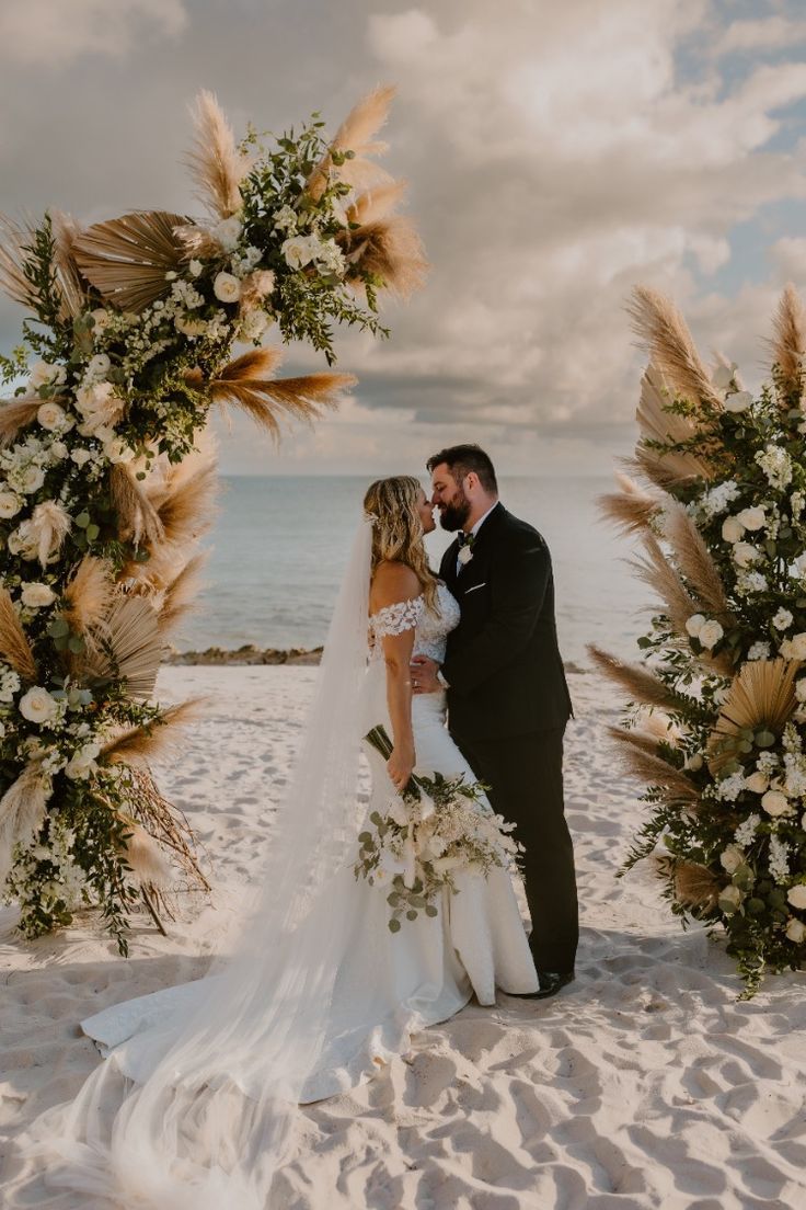 a bride and groom standing under an arch made of pamodia flowers on the beach