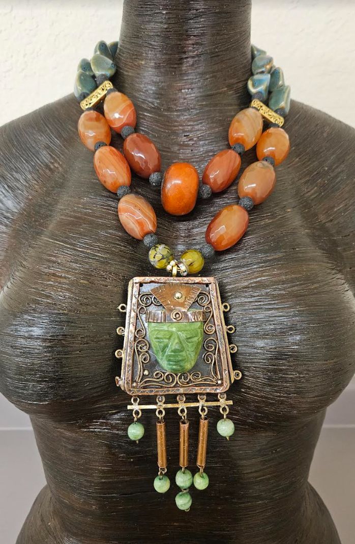 The 5" x 2.5" exotic copper and gemstone Aztec beaded pendant on this piece is authentic to Mexico, but I cannot recall what it is called. A green gemstone is carved into what looks like a Chieftain's face with a copper headdress. The pendant is ornate featuring soldered scrolls and 5 beaded dangles. The adjustable beaded necklace consists of burnt-orange faceted agate beads, black lava spacers, ornate gold tone metal strand spacers, olivine green porcelain beads and coral tubes. Two gold tone aurora borealis rhinestones flank the pendant along with yellow-green agates. This bold and chunky necklace adjusts from 18-21" and is finished with gold tone hardware, an xl fancy lobster claw clasp and a 3" extender chain. One Moroccan resin focal bead is centered on the inner strand. From the Atel Luxury Traditional Beads For Statement Piece, Luxury Southwestern Necklace With Large Stone, Statement Necklace Mexican, Artisan Copper Necklaces With Gemstone Beads, Artisan Copper Necklace With Gemstone Beads, Artisan Beaded Necklace With Large Pendant, Artisan Bronze Jewelry With Polished Beads, Artisan Bronze Beaded Necklaces With Large Beads, Artisan Jade Beaded Necklaces With Gemstone Beads