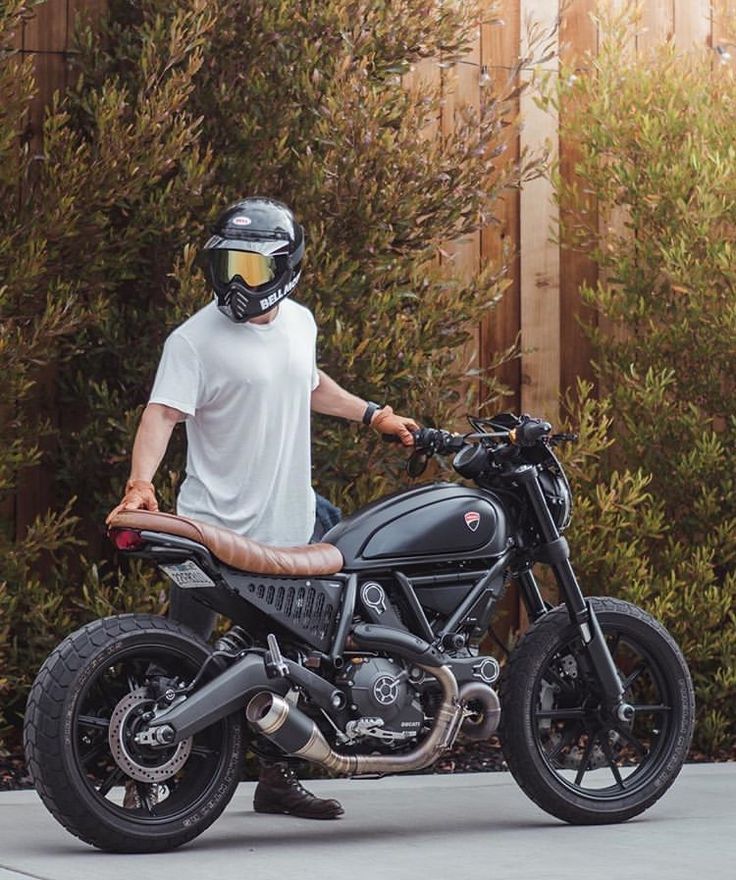 a man in white shirt and black helmet standing next to a motorcycle on street with bushes behind him