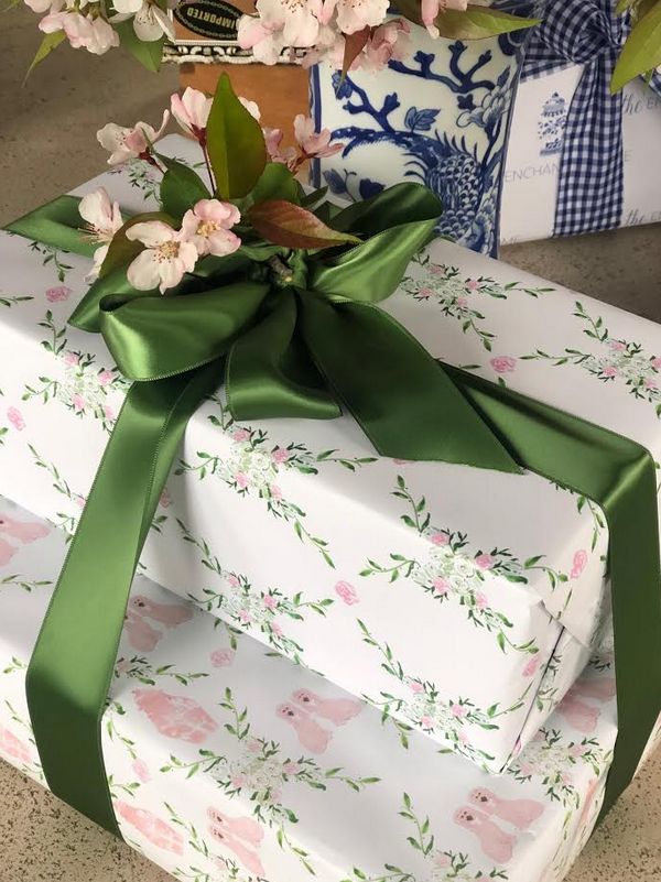 a present wrapped in white paper with green ribbon and pink flowers on the top, sitting next to a vase