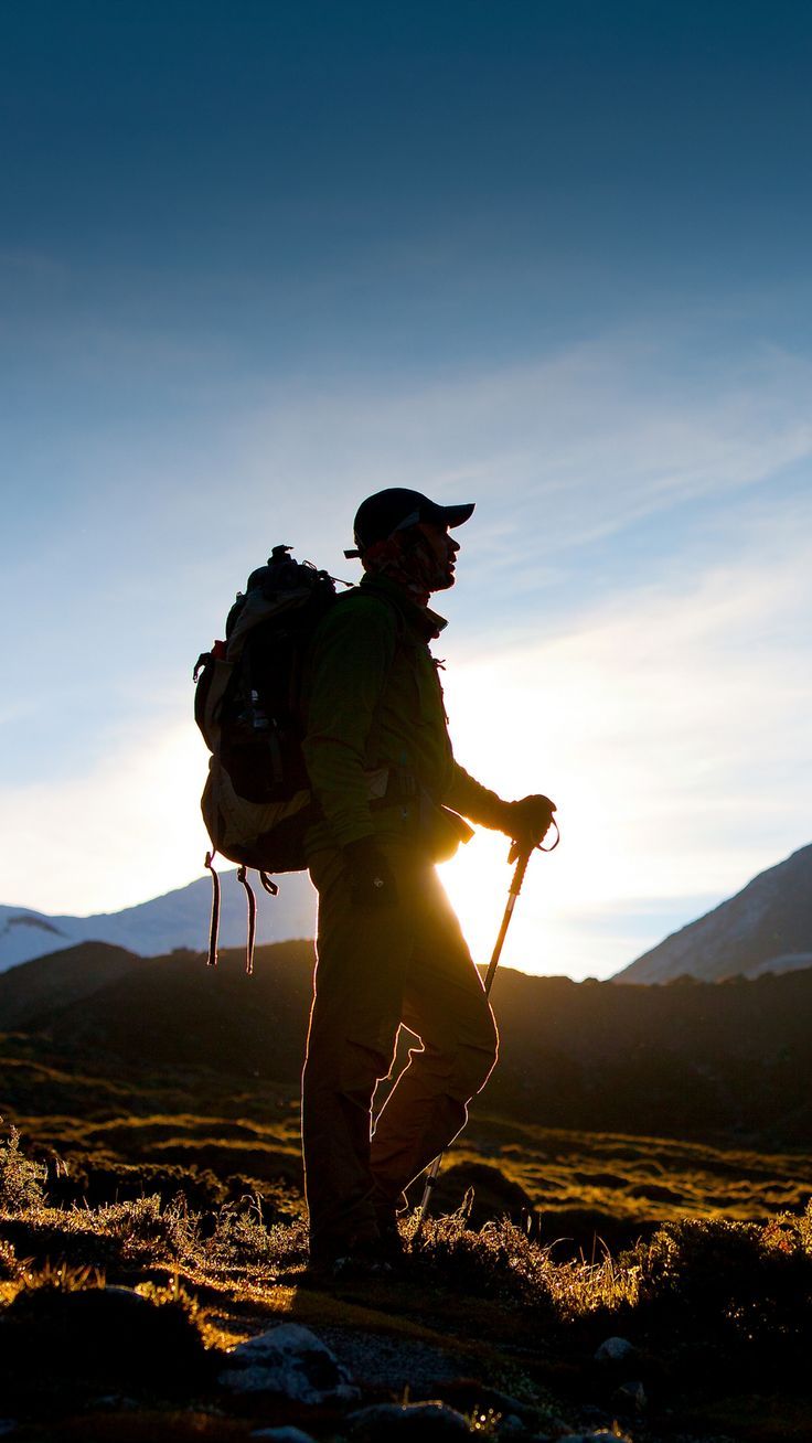 a man with a backpack walking across a grass covered field at sundown in the mountains