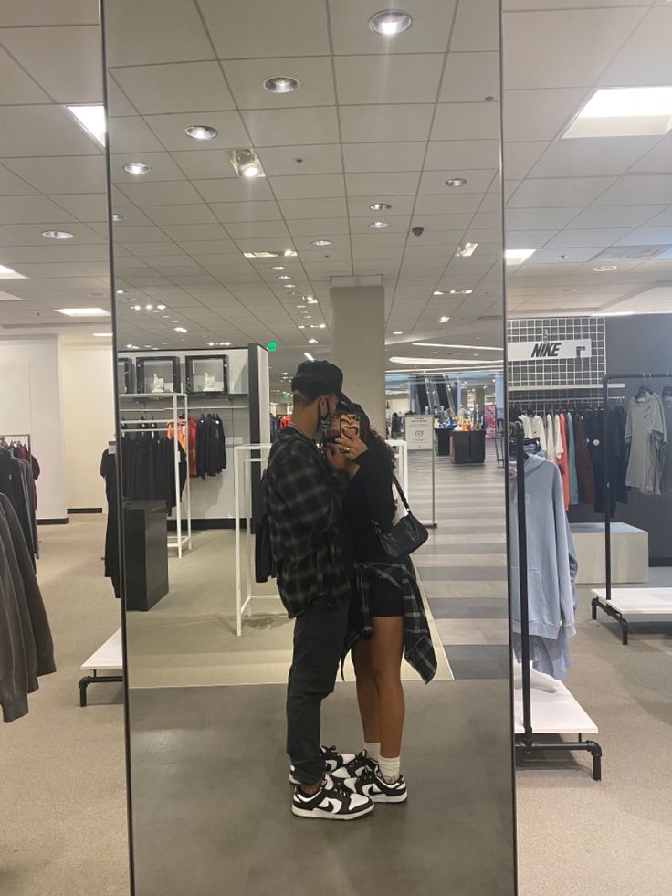 a man and woman taking a selfie in front of a mirrored wall with clothes on display