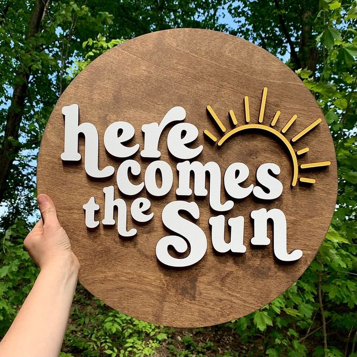 a person holding up a wooden sign that says here comes the sun in front of some trees