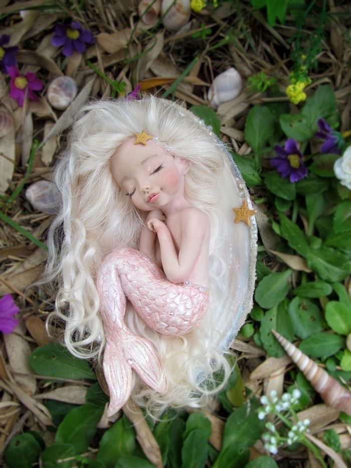 a mermaid doll laying on top of a bed of grass next to flowers and seashells