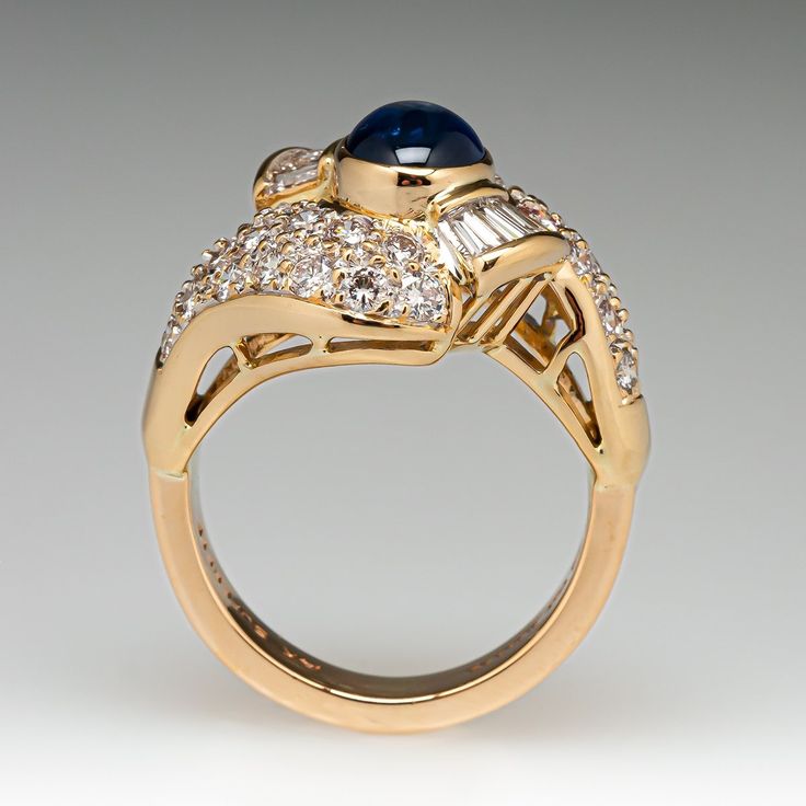 This elegant ring is centered with one (1), bezel set, oval cabochon cut natural sapphire. The sapphire is bordered with eight (8), channel set, tapered baguette cut diamonds and thirty-two (32), bead set, round brilliant cut diamonds. The ring measures 17.0mm at the top, rises 9.7mm above the finger, tapering to 3.8mm wide and 2.0mm thick at the base of the shank. This ring is currently a size 6. Elegant Gold Sapphire Ring With Oval Cabochon, Elegant Sapphire Diamond Ring Channel Set, Formal Domed Cabochon Sapphire Ring, Luxury Yellow Gold Sapphire Ring Channel Set, Elegant Domed Sapphire Ring In Yellow Gold, Elegant Domed Yellow Gold Sapphire Ring, Luxury Oval Sapphire Ring With Polished Finish, Formal Baguette Cut Sapphire Ring Channel Set, Formal Domed Blue Sapphire Ring