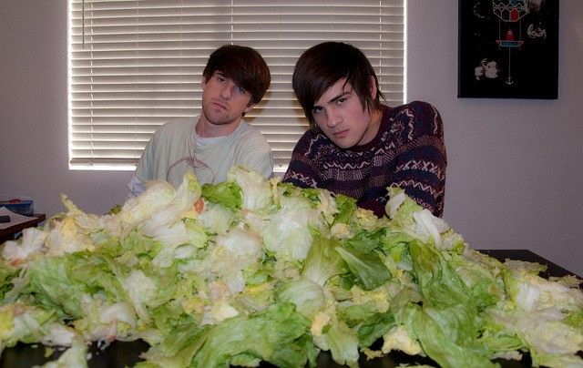 two young men sitting in front of a pile of lettuce on a table