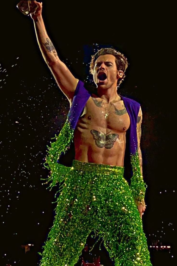 a shirtless man dressed in green and purple is performing on stage with his hands up