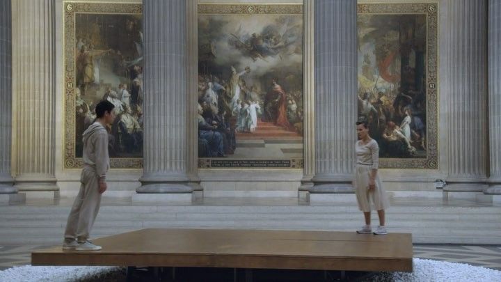 two people standing on a platform in front of large paintings and columns, with one person looking at the ground