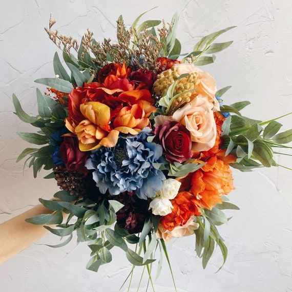 an orange and blue bouquet with greenery on the top is being held by a person