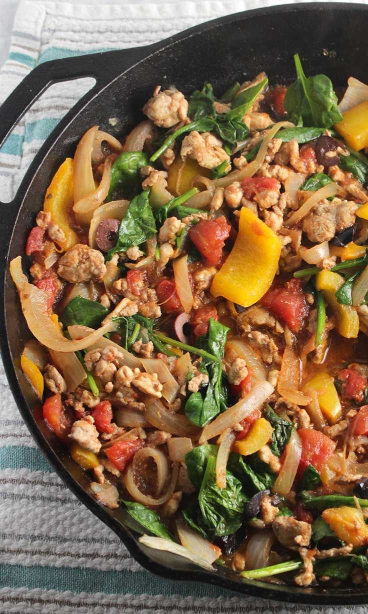 a skillet filled with vegetables and meat on top of a striped table cloth next to a wooden spoon