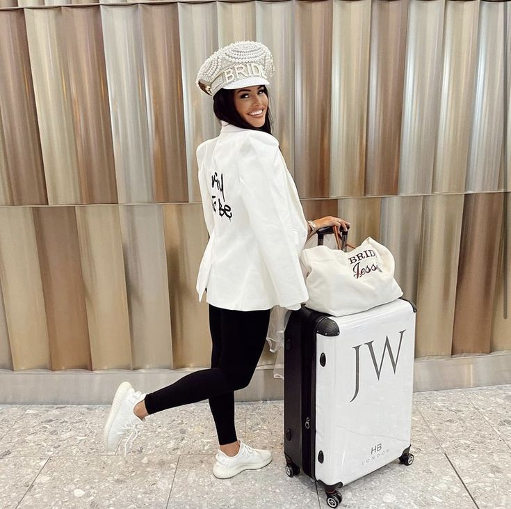 a woman in white jacket and hat pulling luggage with bag next to wall behind her