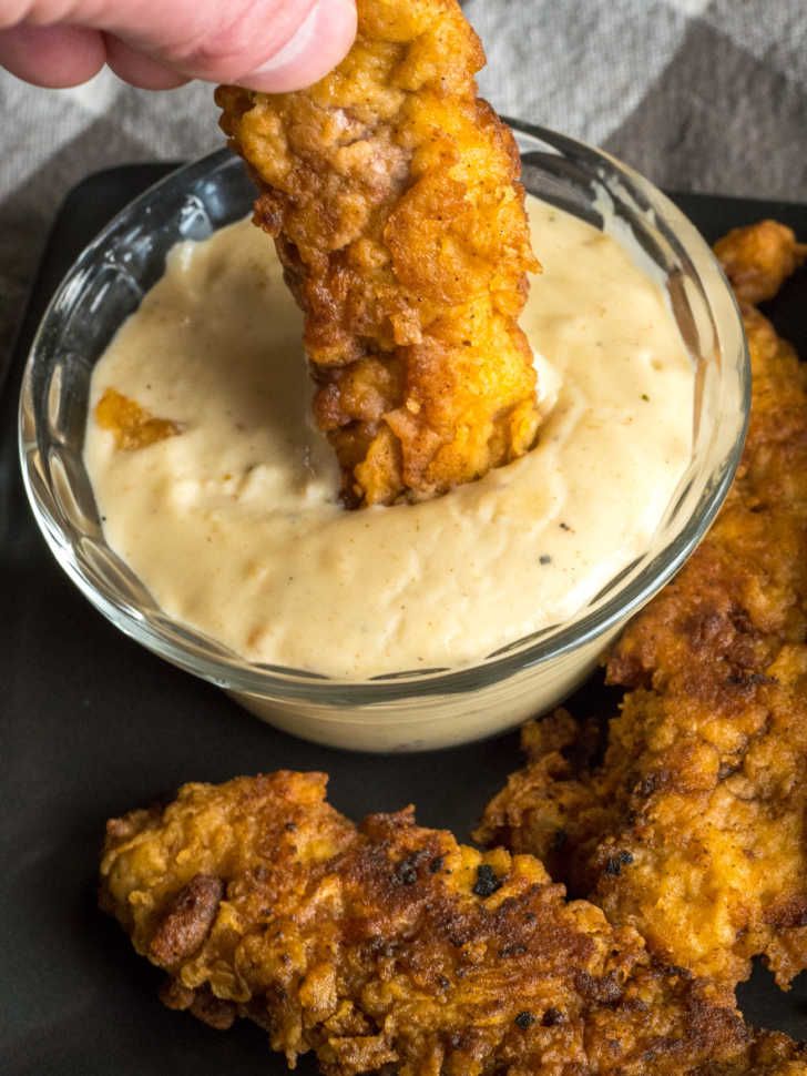 a person dipping something into a bowl with sauce in it and chicken wings on the side