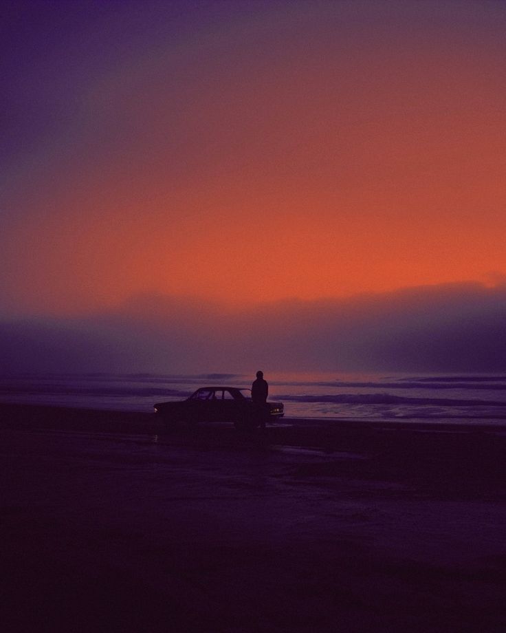 a person standing on the beach at sunset with a car parked in front of them