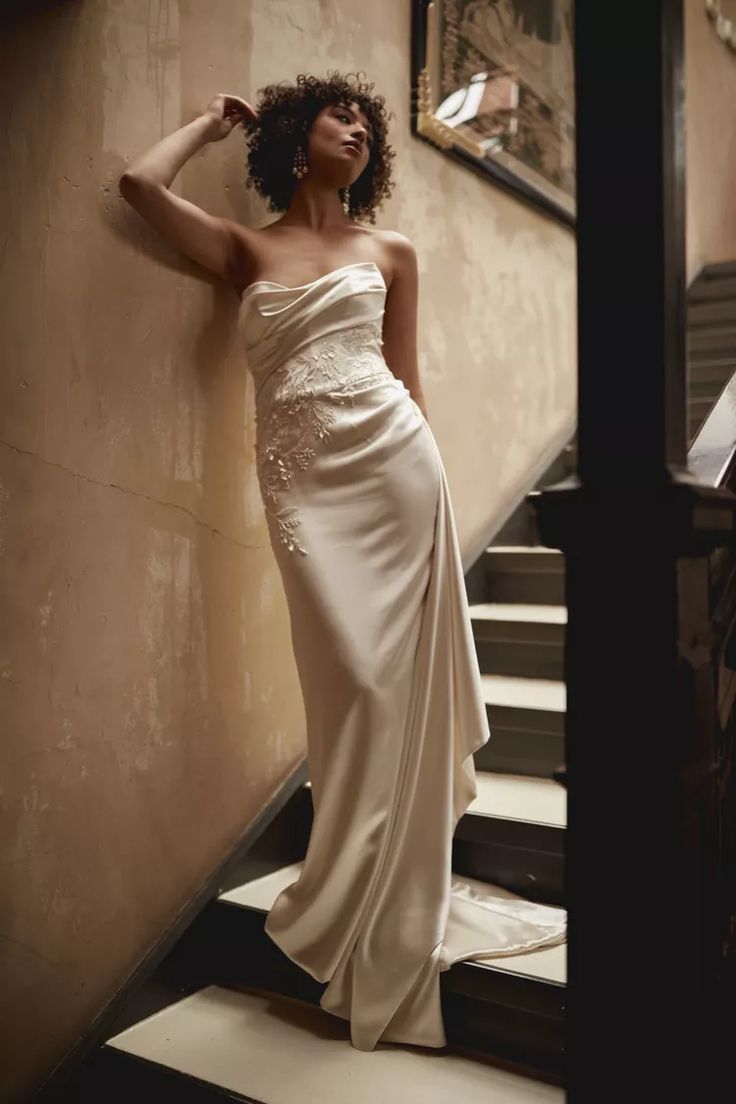 a woman in a white dress leaning against a wall next to some stairs with her hand on her head