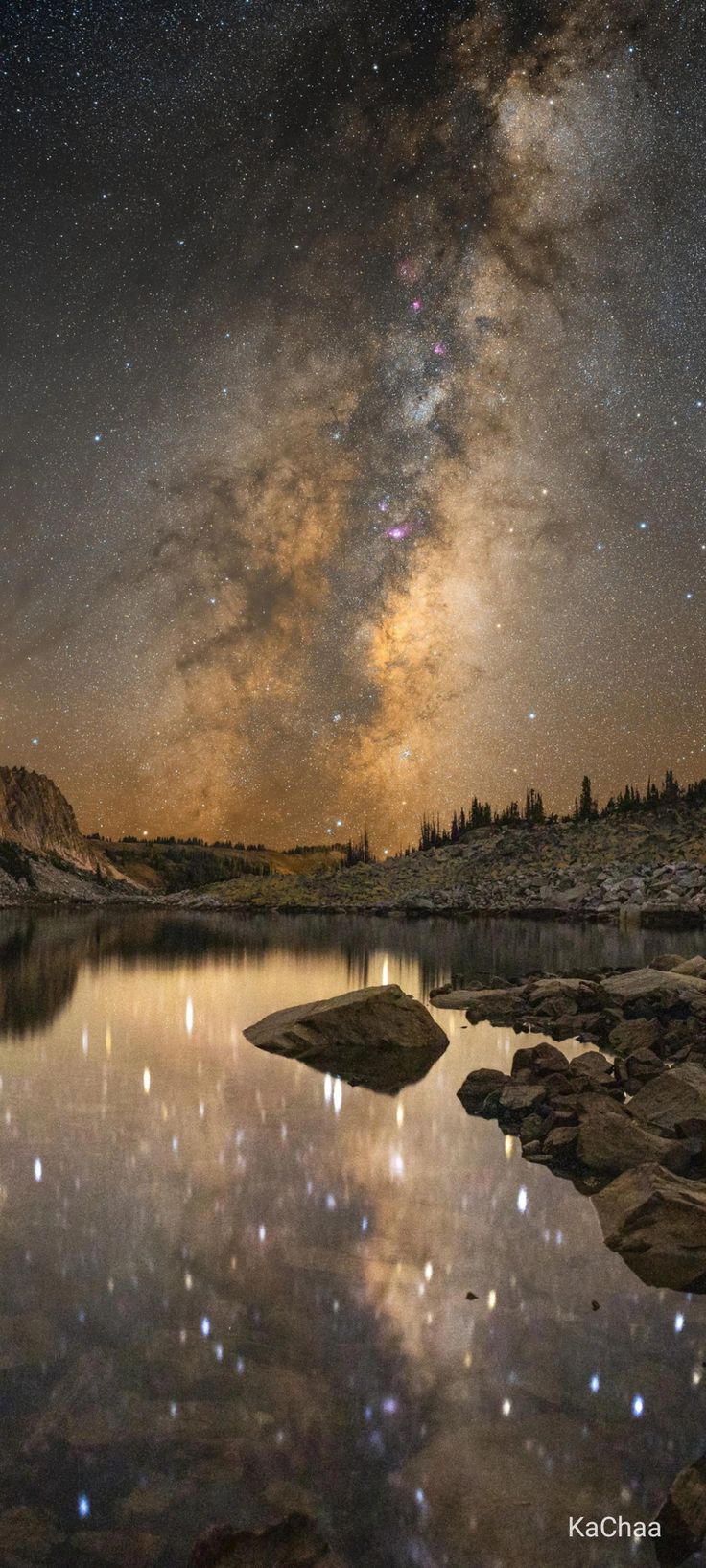 the night sky with stars and clouds reflected in still water on a rocky shore near a mountain lake