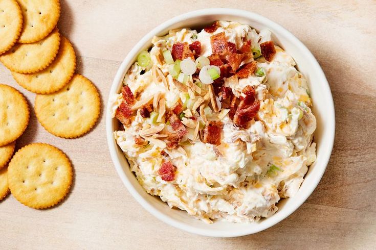 a bowl of potato salad next to crackers on a wooden table with cheese and bacon