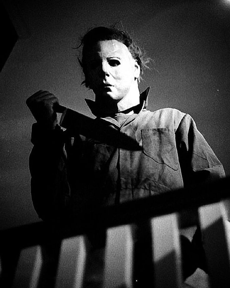 a black and white photo of a man in a scary mask holding a knife up to his face