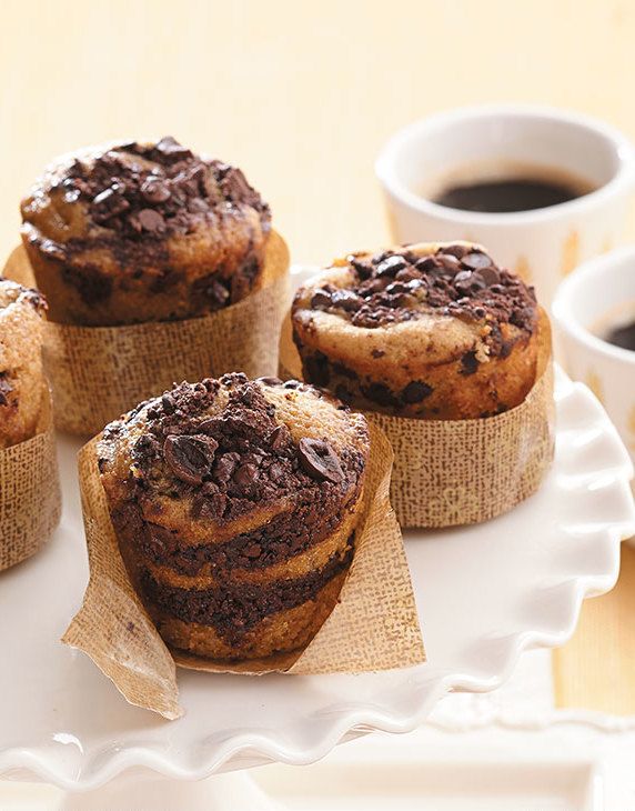 four chocolate chip muffins on a white plate with coffee and saucers in the background