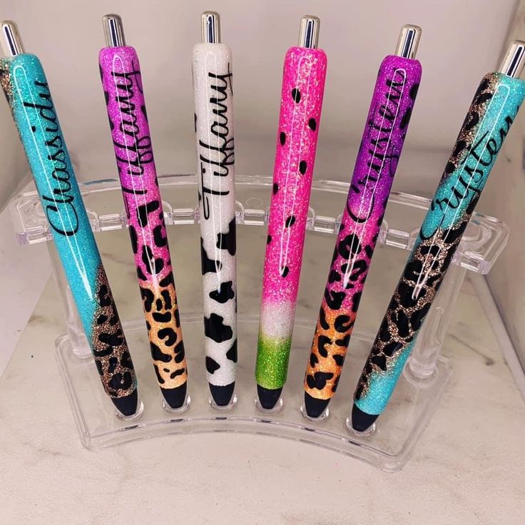 six pens are lined up in a clear holder on a counter top, with different designs and colors