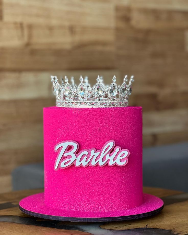 a pink hat with a tiara on top that says barbie in white letters and is sitting on a wooden table