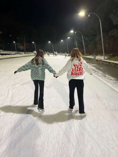 two girls are holding hands while skating on an ice rink at night with street lights in the background