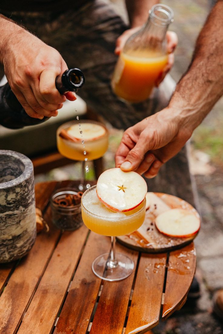 two people are pouring drinks into glasses on a table with apples and other food items