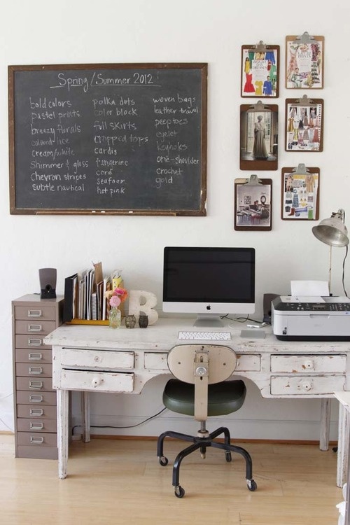 a desk with a computer on it in front of a chalkboard and several pictures