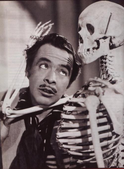 a man holding a skeleton in his right hand and looking at the camera with an evil look on his face