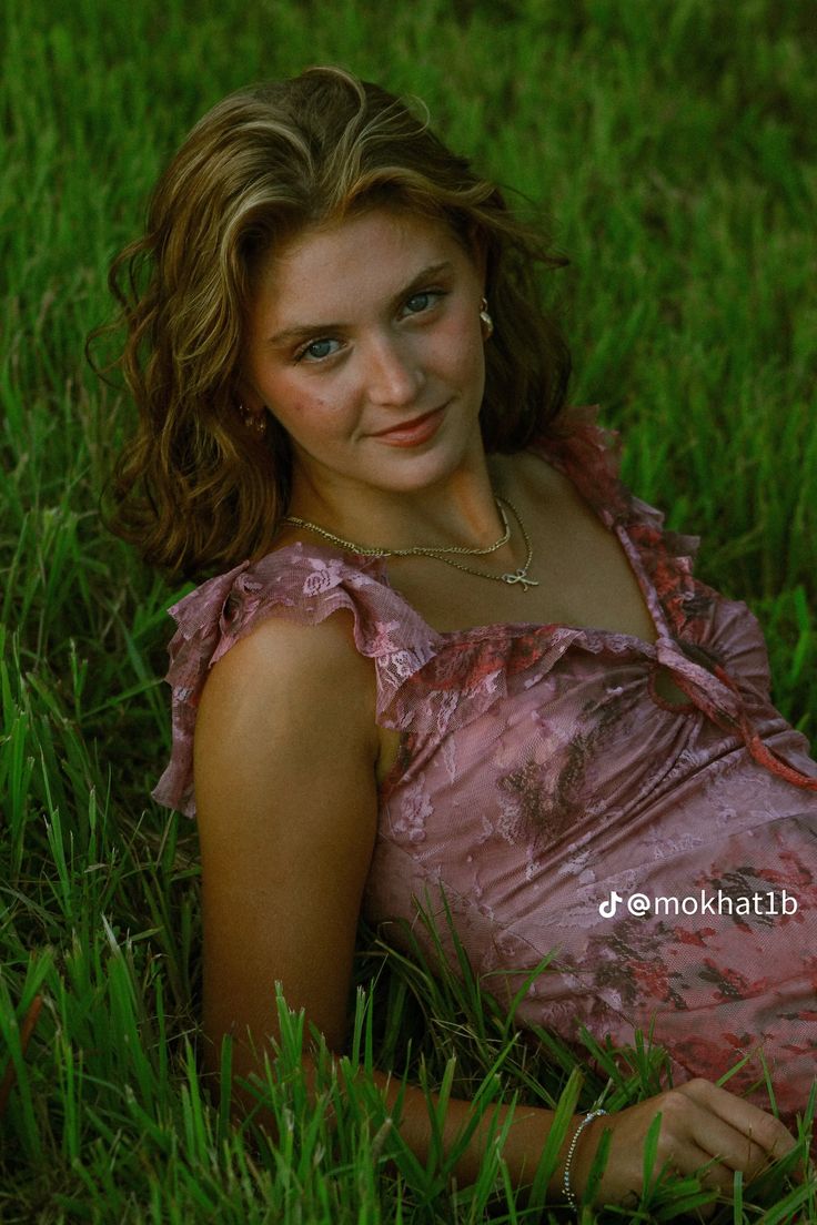 a woman laying in the grass wearing a pink dress and smiling at the camera with her hand on her hip