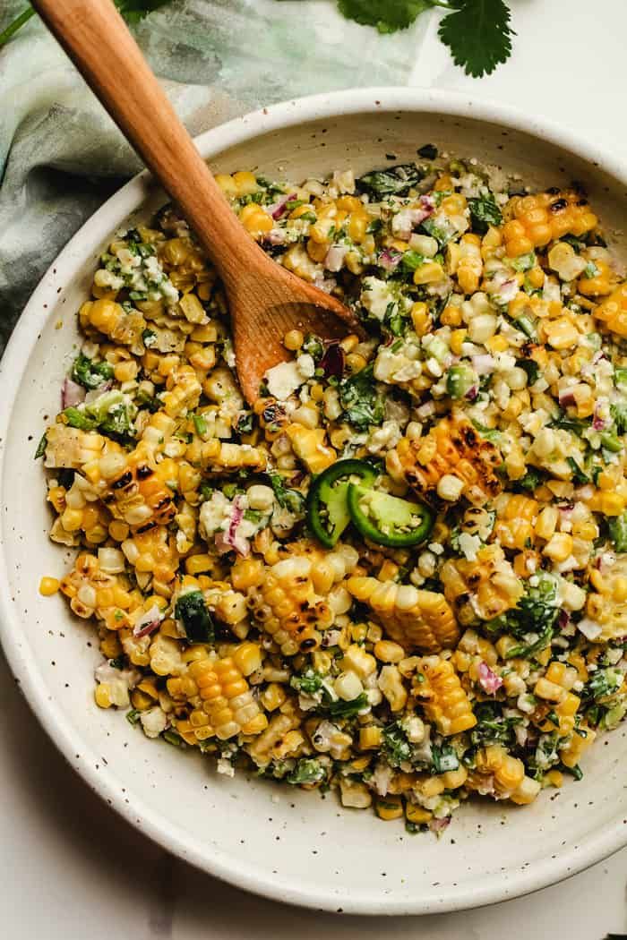 a bowl filled with corn and vegetables on top of a white table next to a wooden spoon