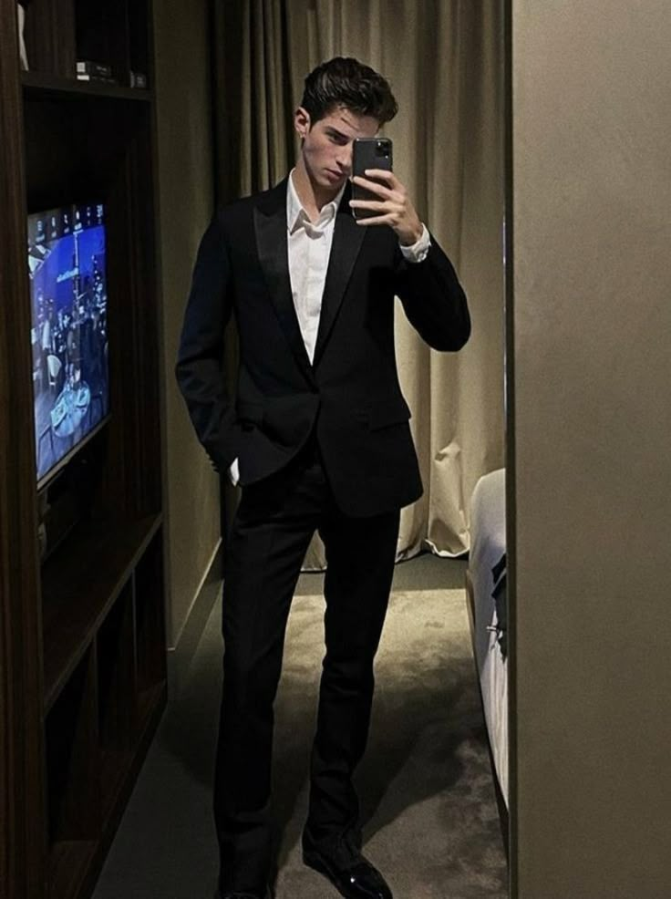 a man in a suit taking a selfie with his cell phone while standing next to a bed