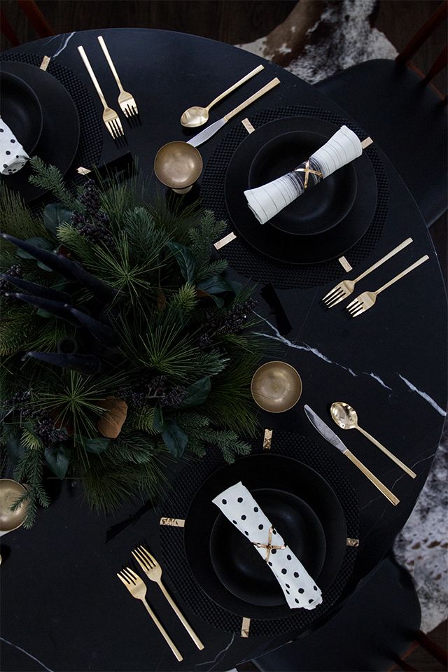 the table is set with black plates and gold cutlery, silverware, and greenery