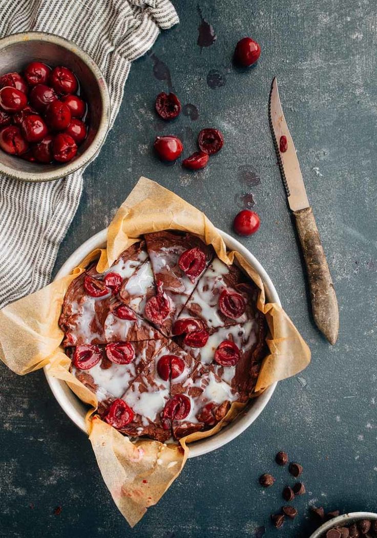 a cherry pie with icing and cherries in bowls next to a knife, fork and bowl of cherries