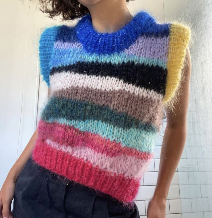 a woman wearing a multicolored sweater and black pants standing in front of a white tiled wall