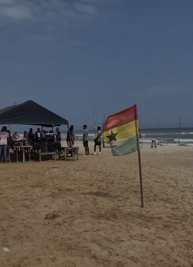 a flag on the beach with people standing and sitting at tables in front of it