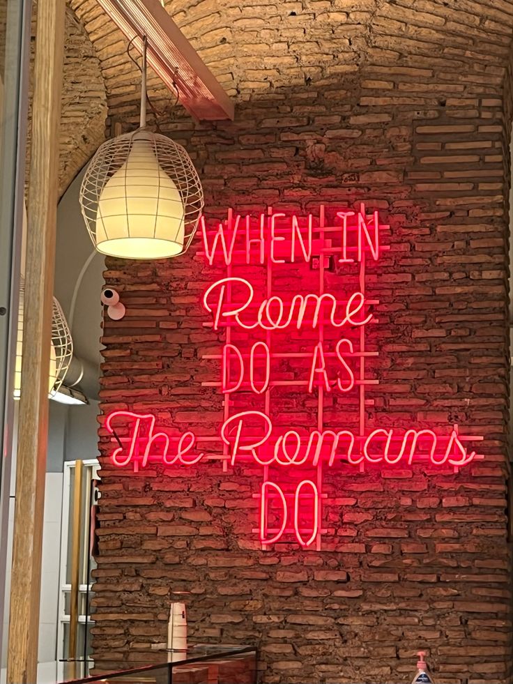 a red neon sign that says when rome does the romans do on a brick wall