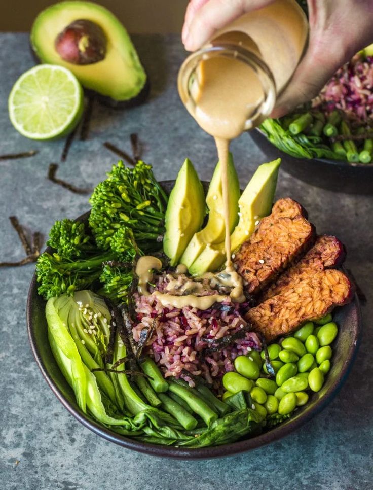 someone pouring dressing onto a plate of food with vegetables and avocado on the side