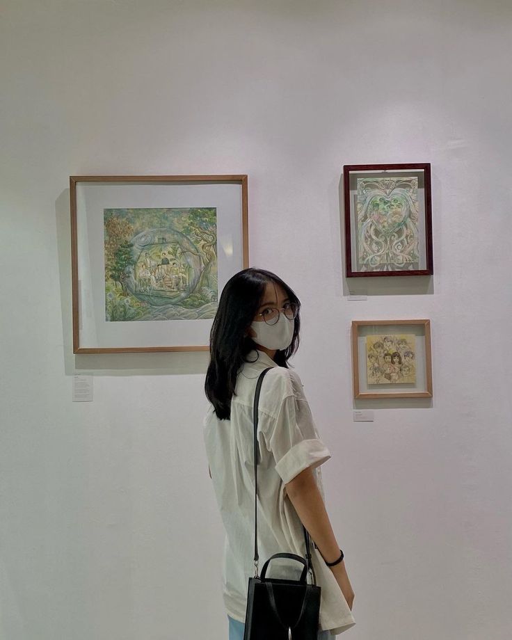 a woman wearing a face mask standing in front of paintings on the wall and holding a black purse