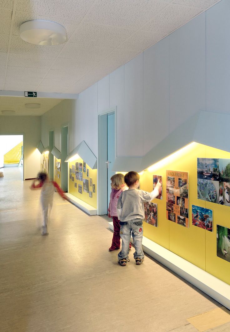 two children are standing in front of a yellow wall with pictures on it and people walking by