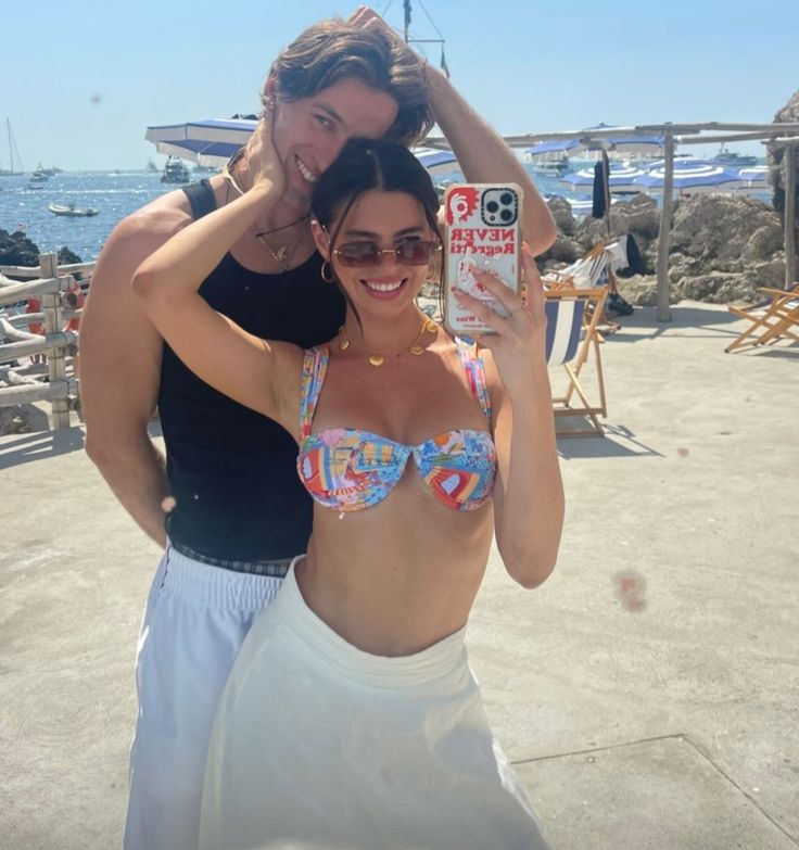 two women in bikinis are posing for a photo on the beach with their cell phone