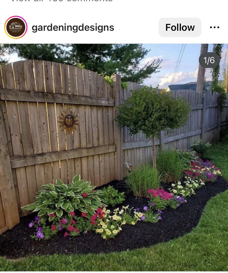 a garden with flowers and plants in the grass next to a wooden fence on a sunny day