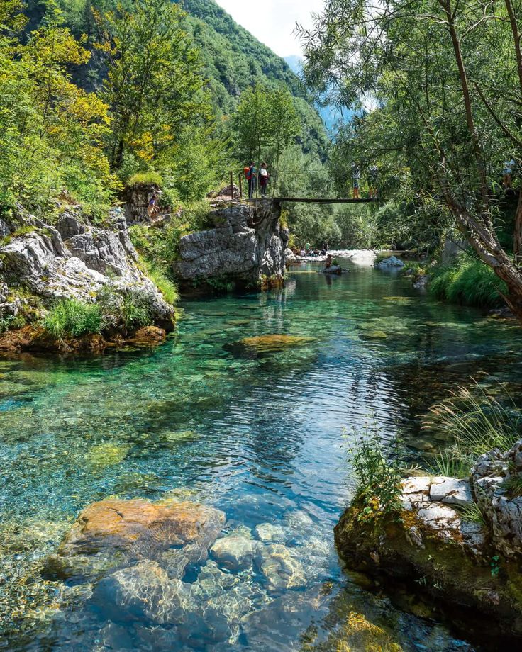 people are standing on a bridge over a river with clear water and green mountains in the background