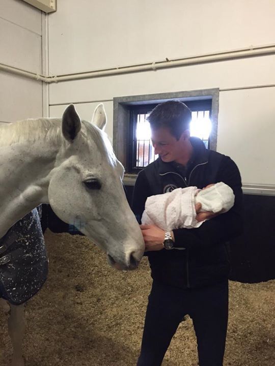 a man holding a baby while standing next to a white horse in an indoor barn